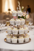 Load image into Gallery viewer, A five-tier silver cupcake stand holding decorated cupcakes with white frosting, white lace-patterned liners, and small pale lavender flower toppers against a warm brown textured background.
